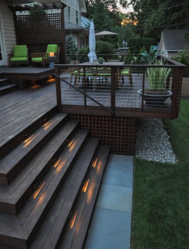 A multi-level wooden deck at dusk, featuring glowing recessed stair lights, green lounge chairs, and a dining area.