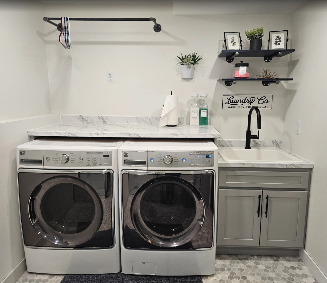 A modern laundry room featuring a washer, dryer, marble countertop, sink, and open shelving on a white wall.