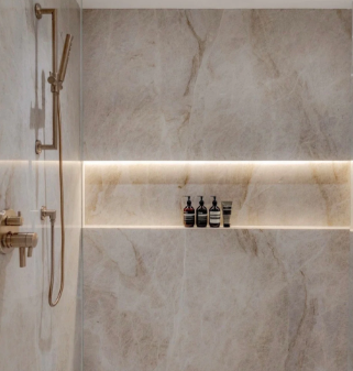 Modern shower with light brown marble tiles, a gold shower fixture, and a recessed shelf illuminated by LED lighting.