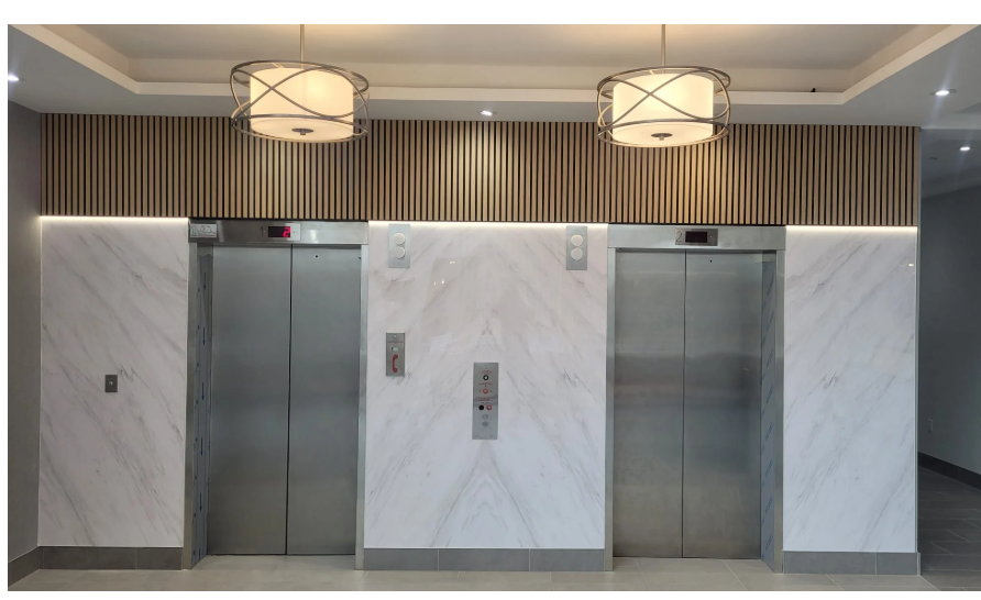 Two stainless steel elevator doors set in a white marble wall with wooden slats above and two pendant lights.