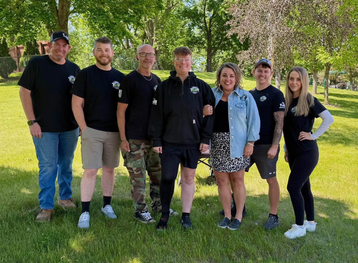 Seven people posing outdoors on a grassy lawn, smiling in casual clothes and black shirts.
