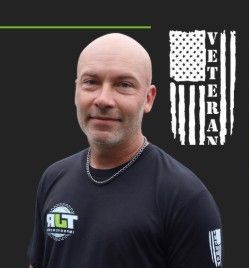 A bald man wearing a black t-shirt is standing in front of an american flag.