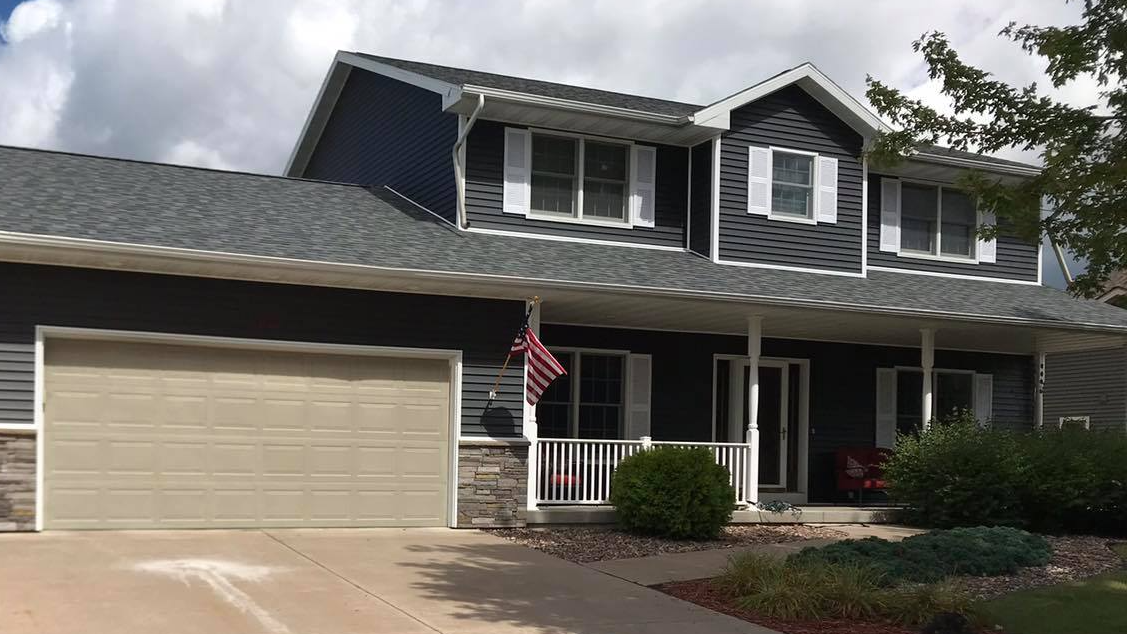 Two-story house with dark blue siding, gray roof, white trim, beige garage door, and a small porch with an American flag.