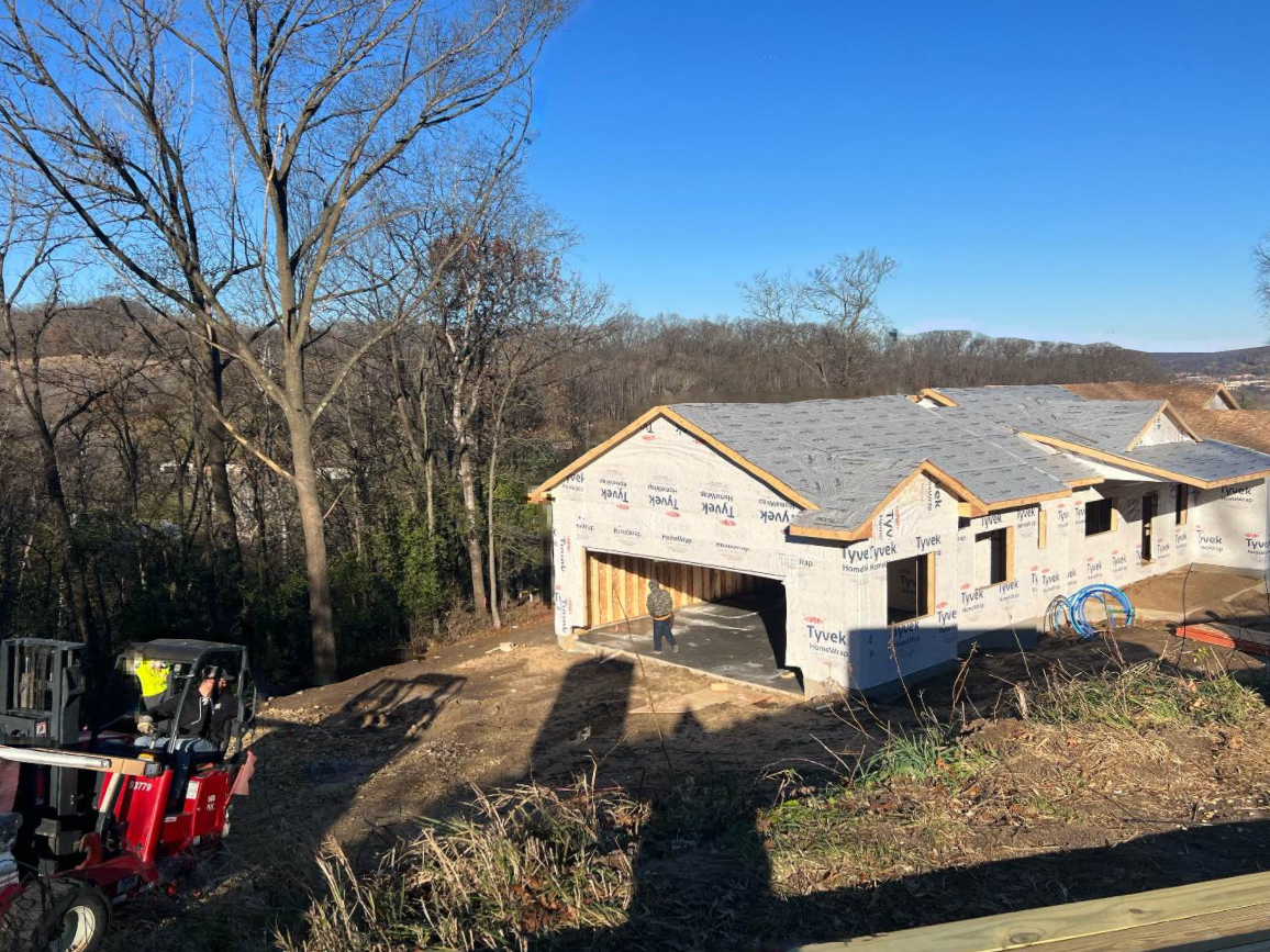 House under construction with a garage, blue sky, and a forklift on a hillside.