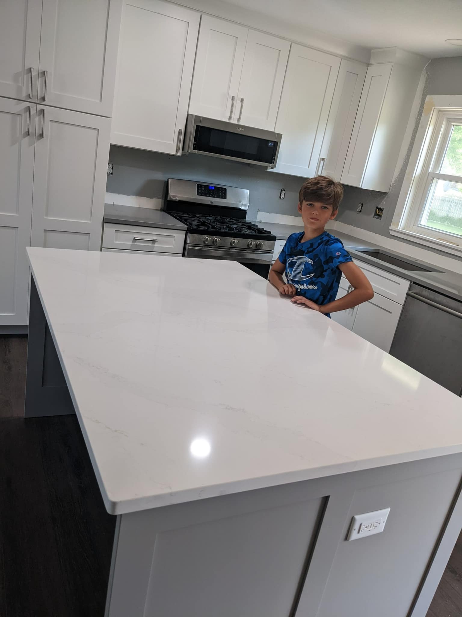 Boy standing by a white quartz kitchen island. Gray cabinets, stove, and dishwasher in background.