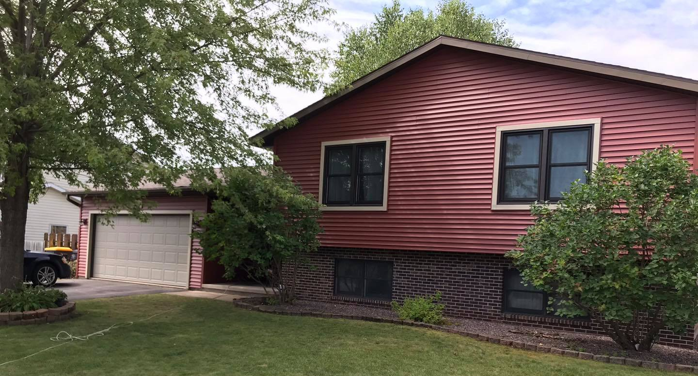 Red house with attached garage, trees, and green lawn. Driveway leads to garage.