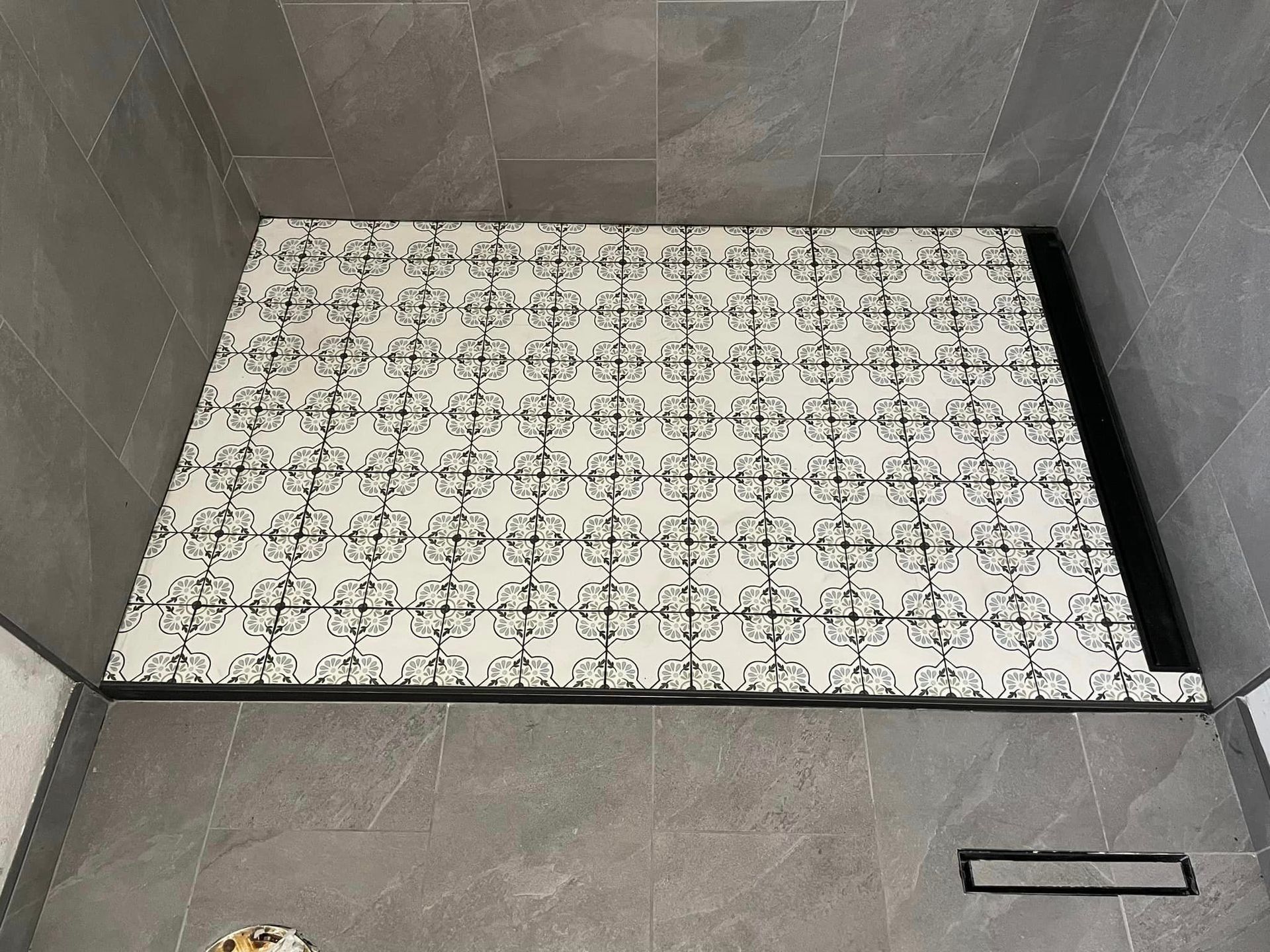 Shower base with a decorative black and white patterned surface, against gray tile walls, and a black linear drain.