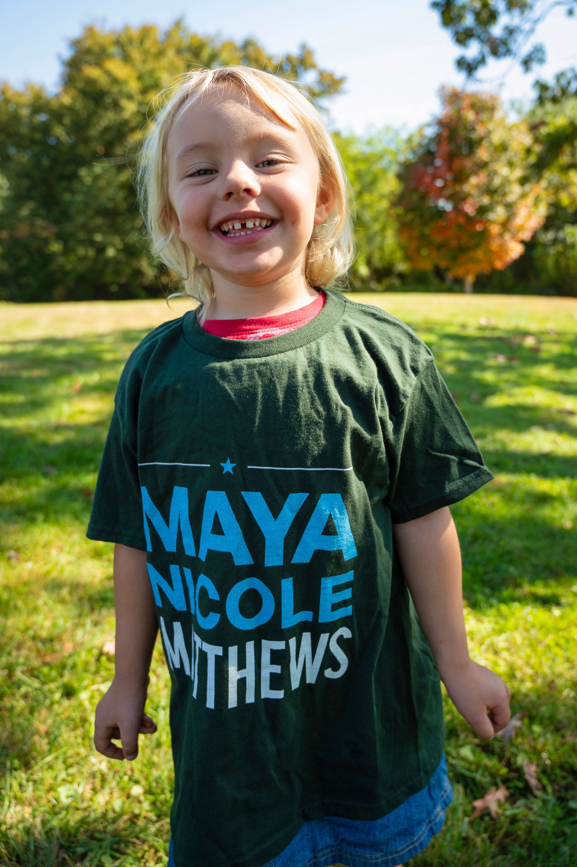 Young child with blond hair smiles, wearing a green t-shirt that says 