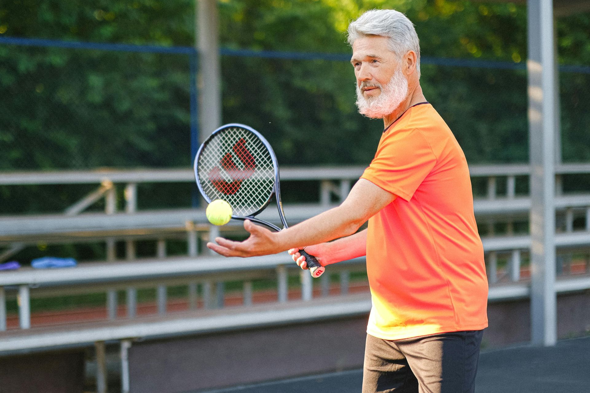 Older man with gray beard playing tennis, tossing ball in air, orange shirt.