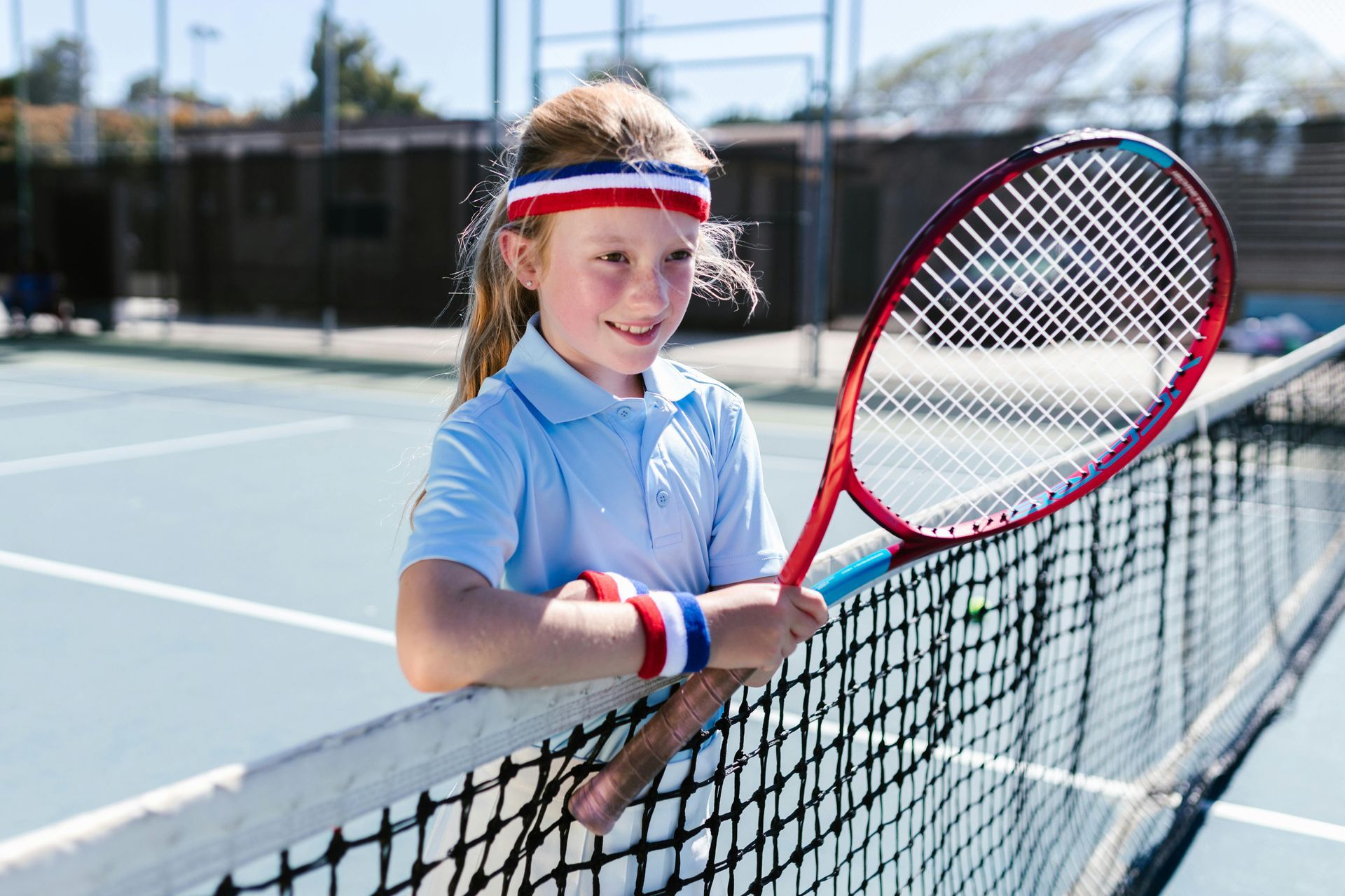 Girl in tennis gear, holding racquet, smiling at net on court.