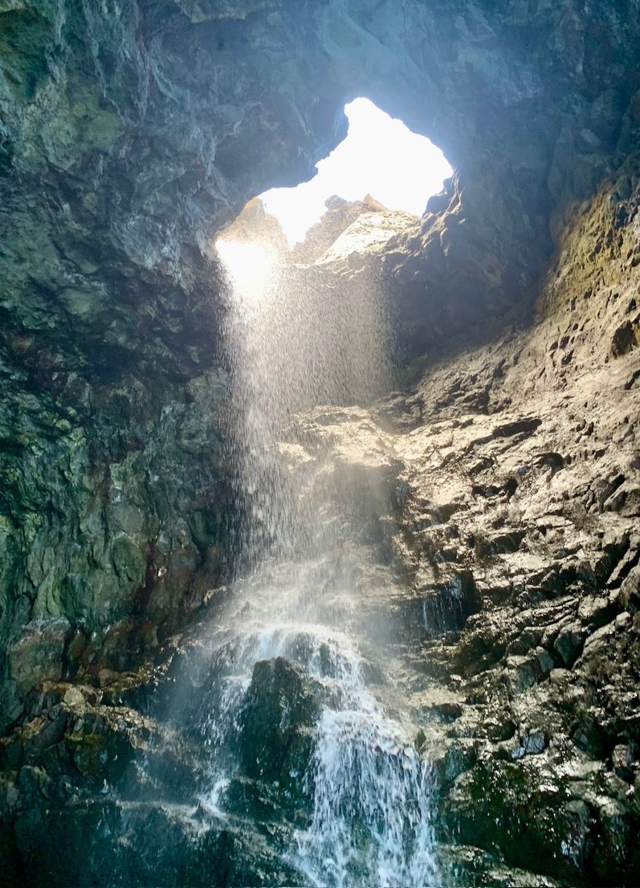 A waterfall in a cave with the sun shining through the opening.