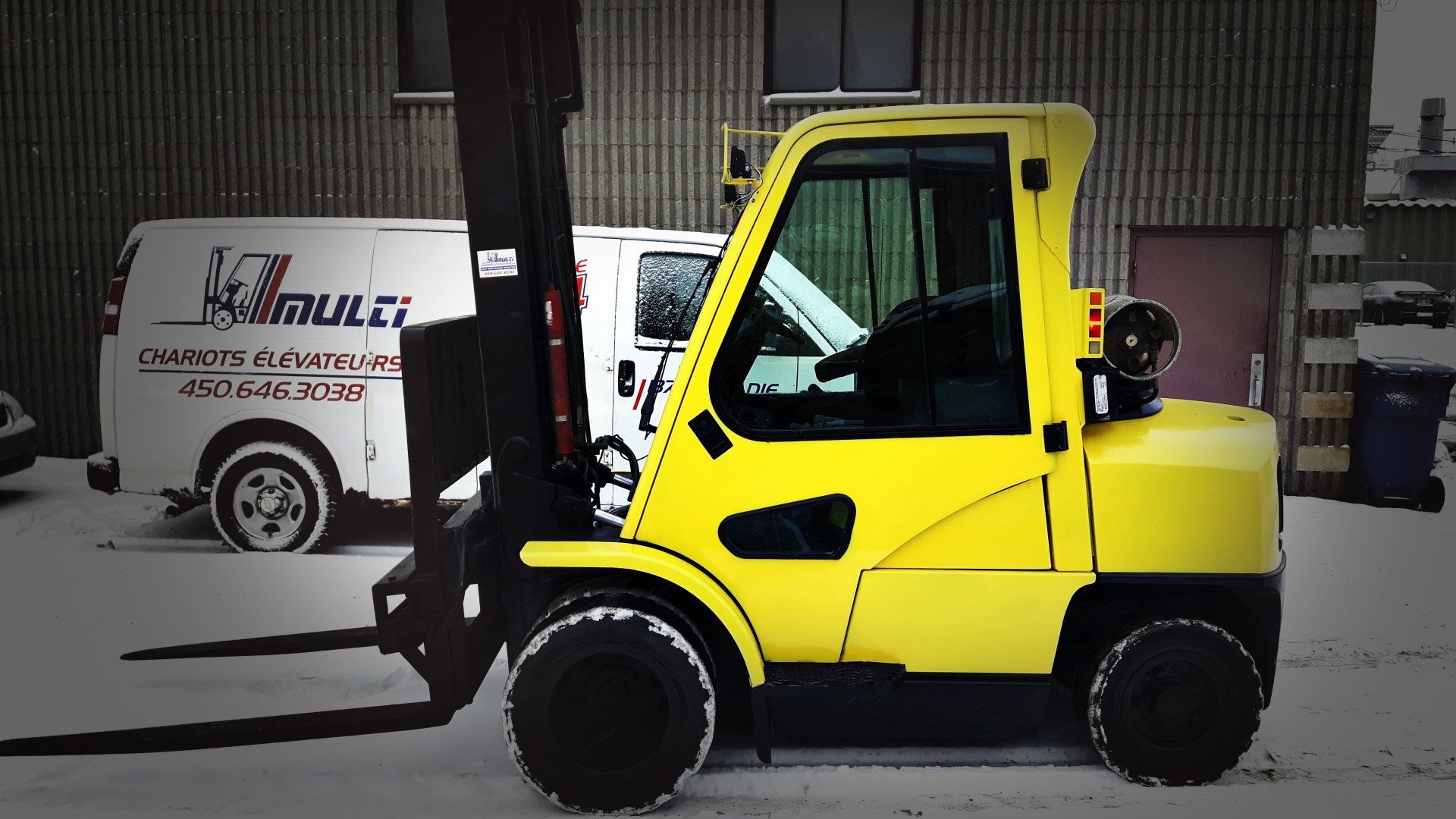 Chariot élévateur jaune sur un sol enneigé, garé près d'une camionnette blanche.