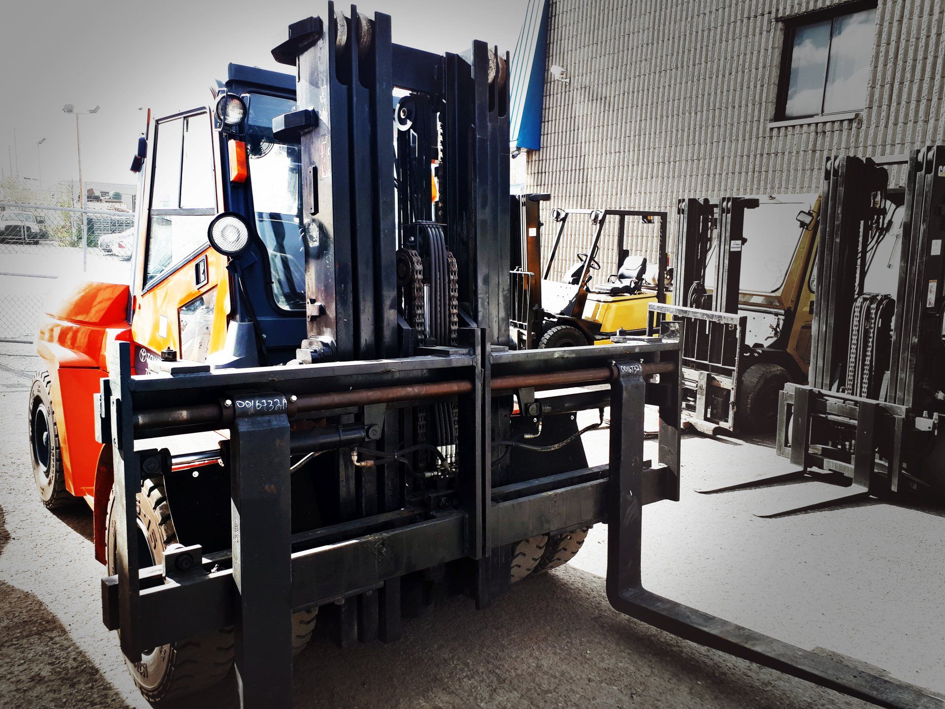 Chariot élévateur orange avec fourches noires garé devant un entrepôt, avec d'autres chariots élévateurs en arrière-plan.