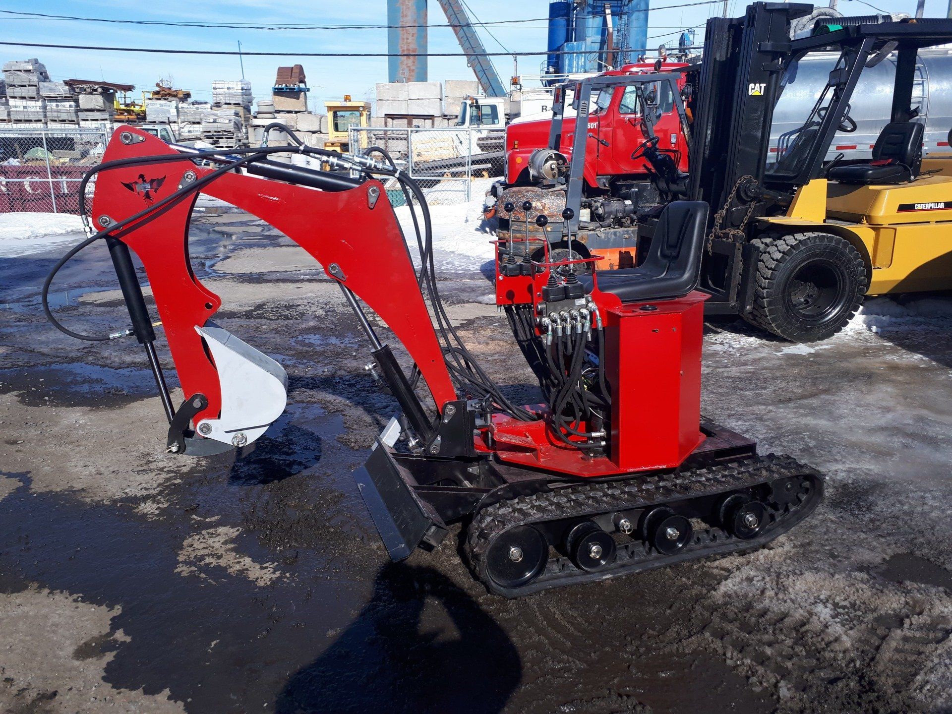 Une mini-pelle rouge sur chenilles, stationnée à l'extérieur sur une surface humide, avec un chariot élévateur jaune en arrière-plan.