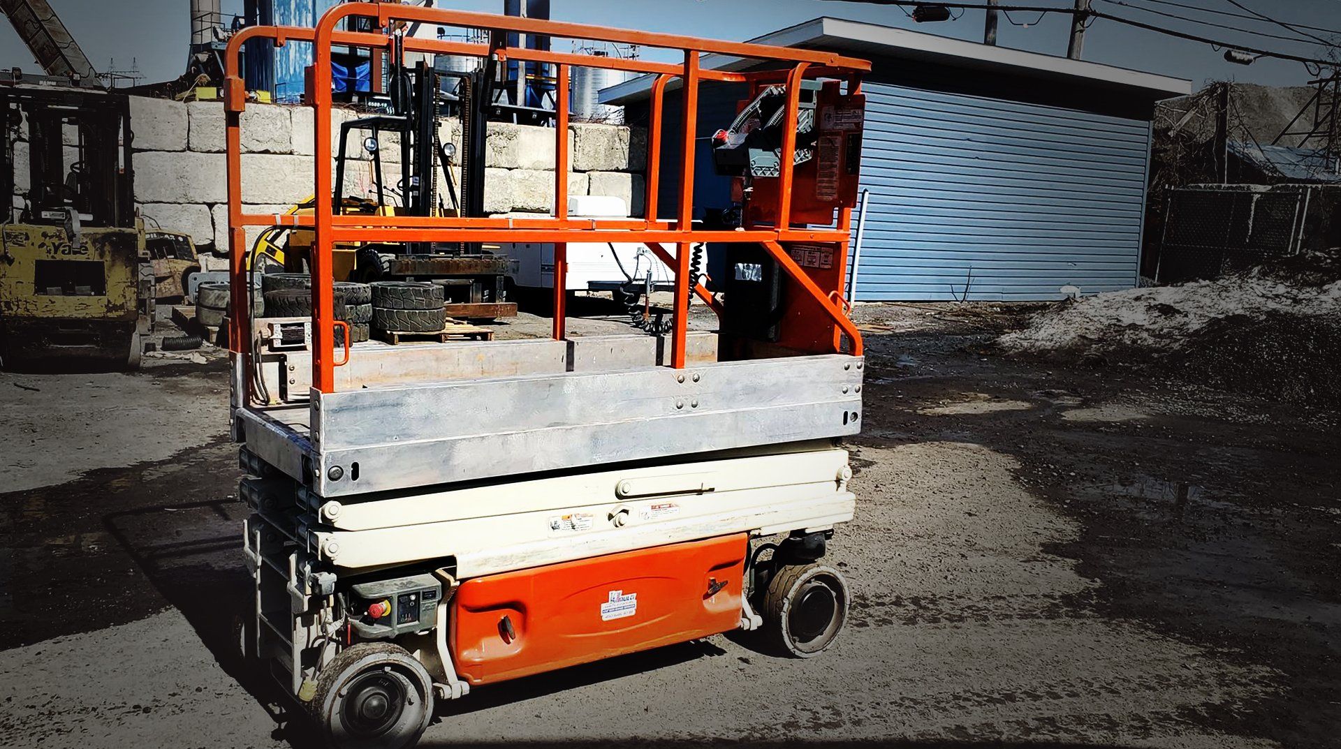Nacelle élévatrice à ciseaux orange et argent sur du gravier, près d'un bâtiment.