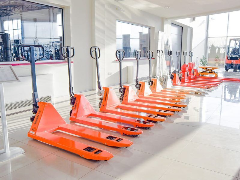 Une rangée de transpalettes orange dans un entrepôt lumineux, avec des fenêtres et un chariot élévateur visibles.