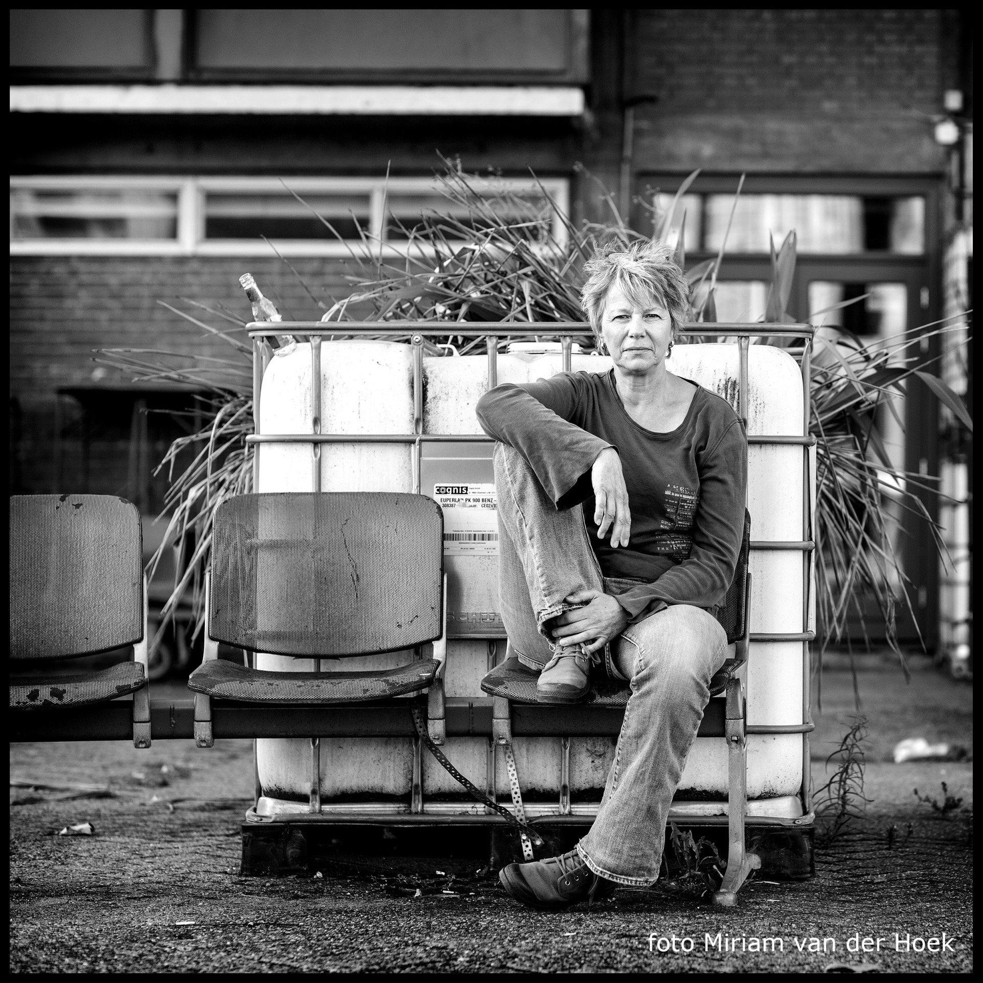 Vrouw zittend op watertanks met stoel, takken, tegen een gebouw in zwart-wit.