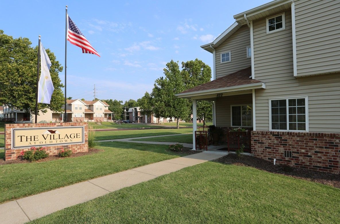 The Village apartments sign, flagpole with American flag, and building with brown brick facade.