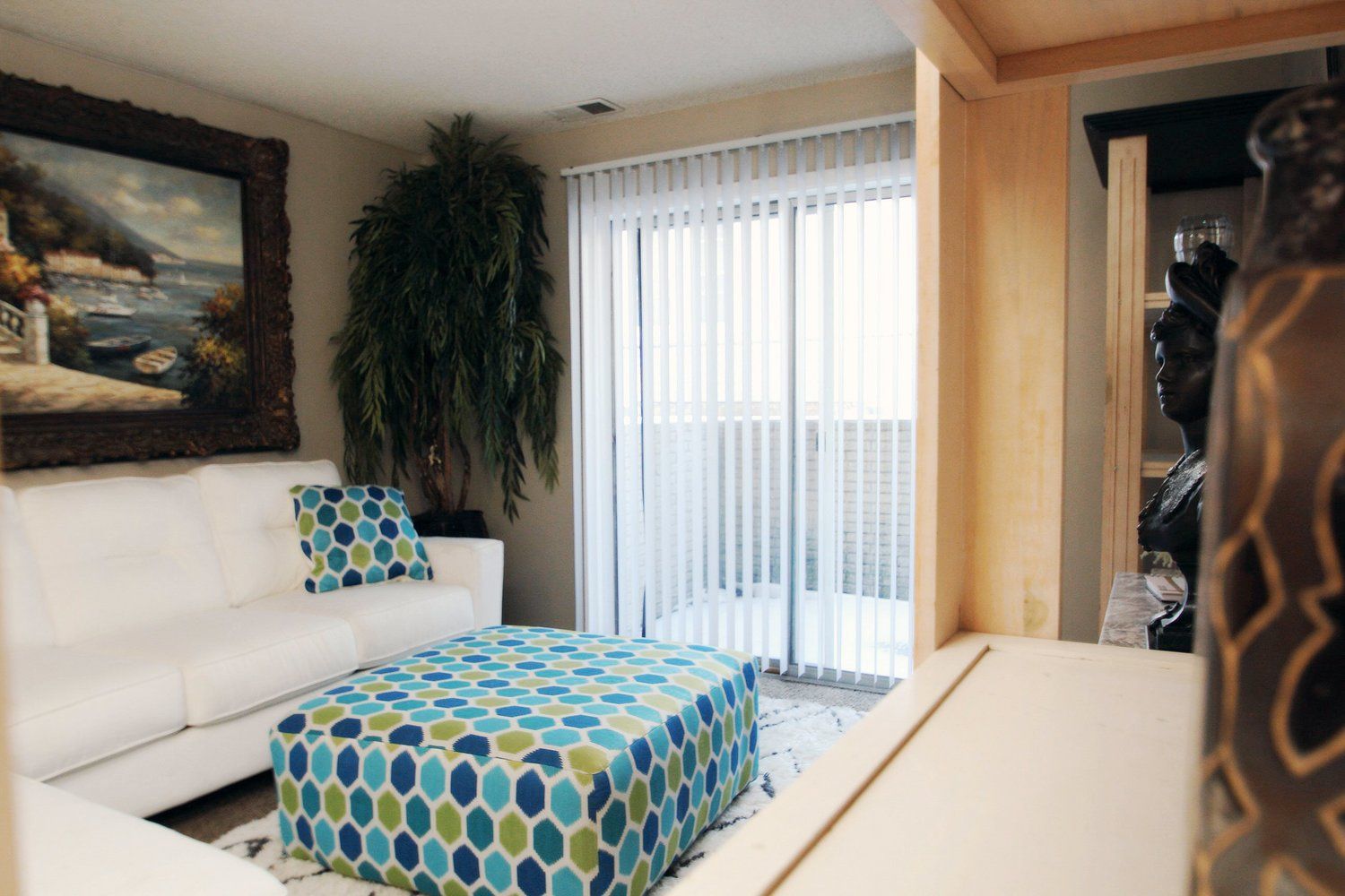 White couch and patterned ottoman in living room with large painting, vertical blinds, and hanging plant.
