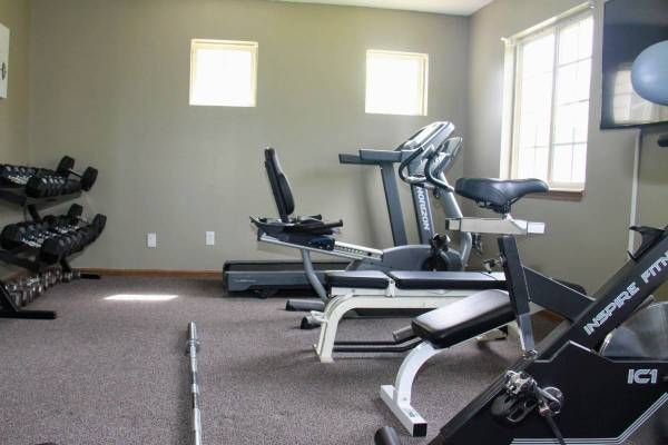 Home gym with exercise equipment: weights, treadmill, recumbent bike, upright bike, and small windows.