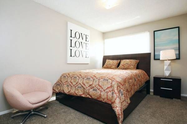 Bedroom with bed, art, pink chair, and nightstand. The walls are light grey, and the bed has a patterned comforter.