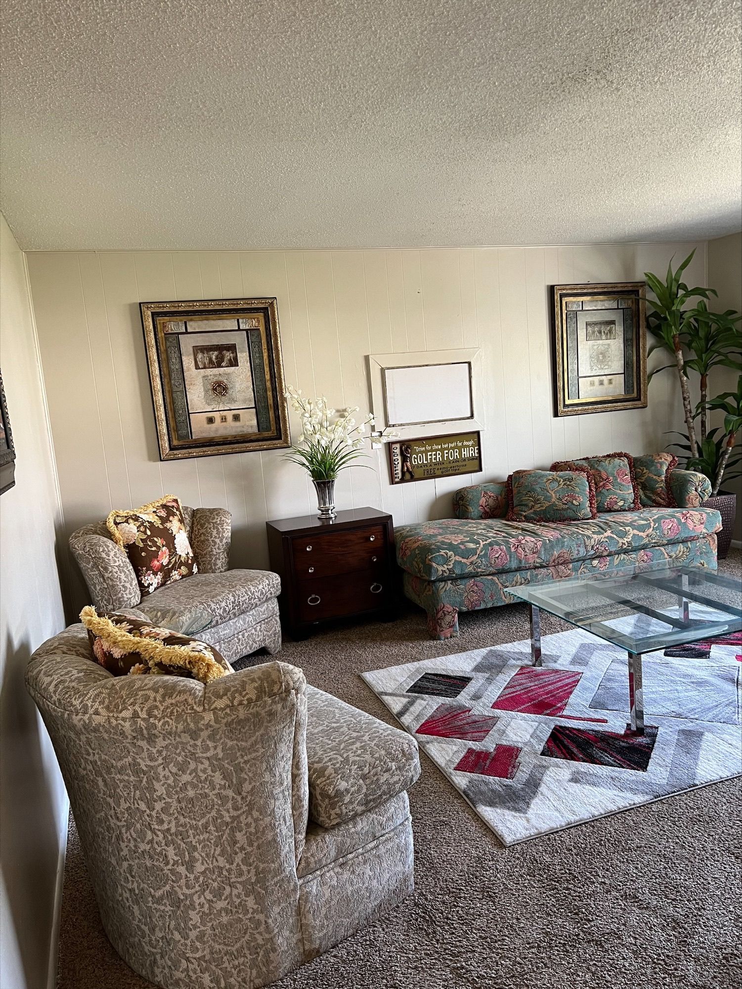 Living room with floral patterned sofa and armchairs, rug, side table, framed artwork, and plants.