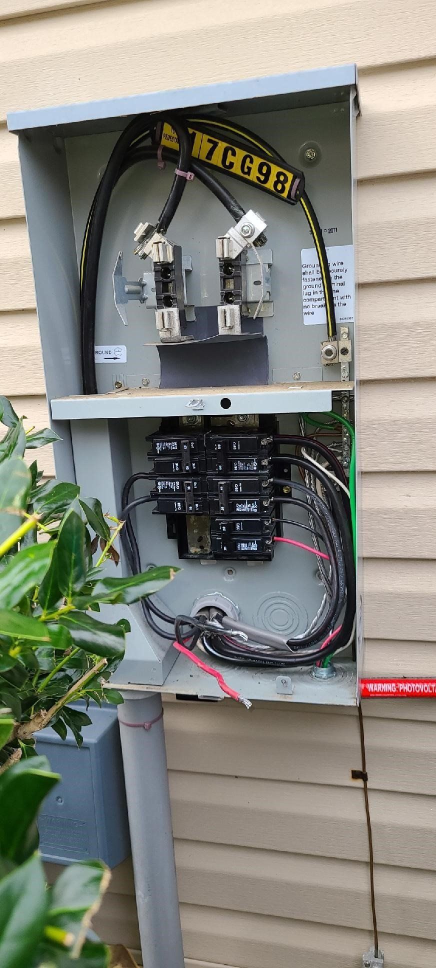 A electrical box is sitting on the side of a house next to a tree.