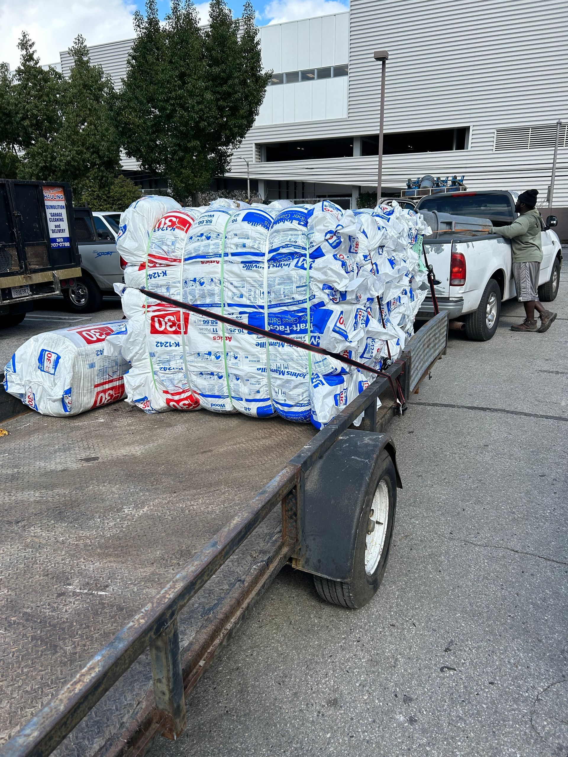 A person stands beside a pickup truck hitched to a trailer loaded with large, white, shrink-wrapped bundles in a lot.