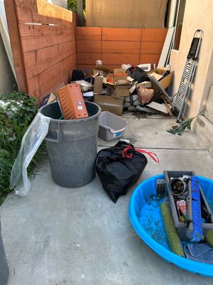 A clutter-filled outdoor area with a grey trash bin, a black bag, a blue plastic tub, and scattered debris by a brick wall.