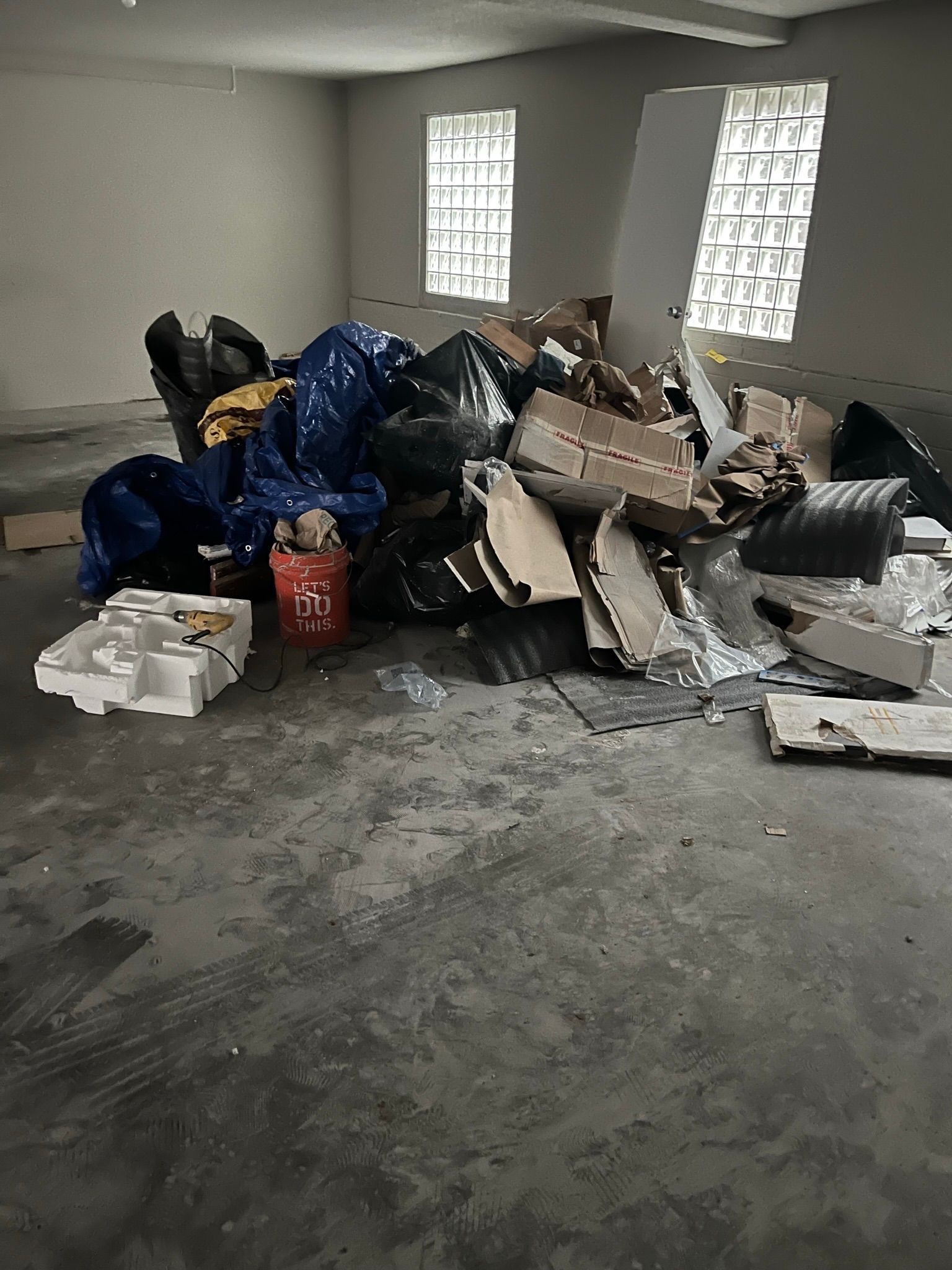 A pile of trash, cardboard boxes, and blue tarps sits in the center of an empty room with glass block windows.