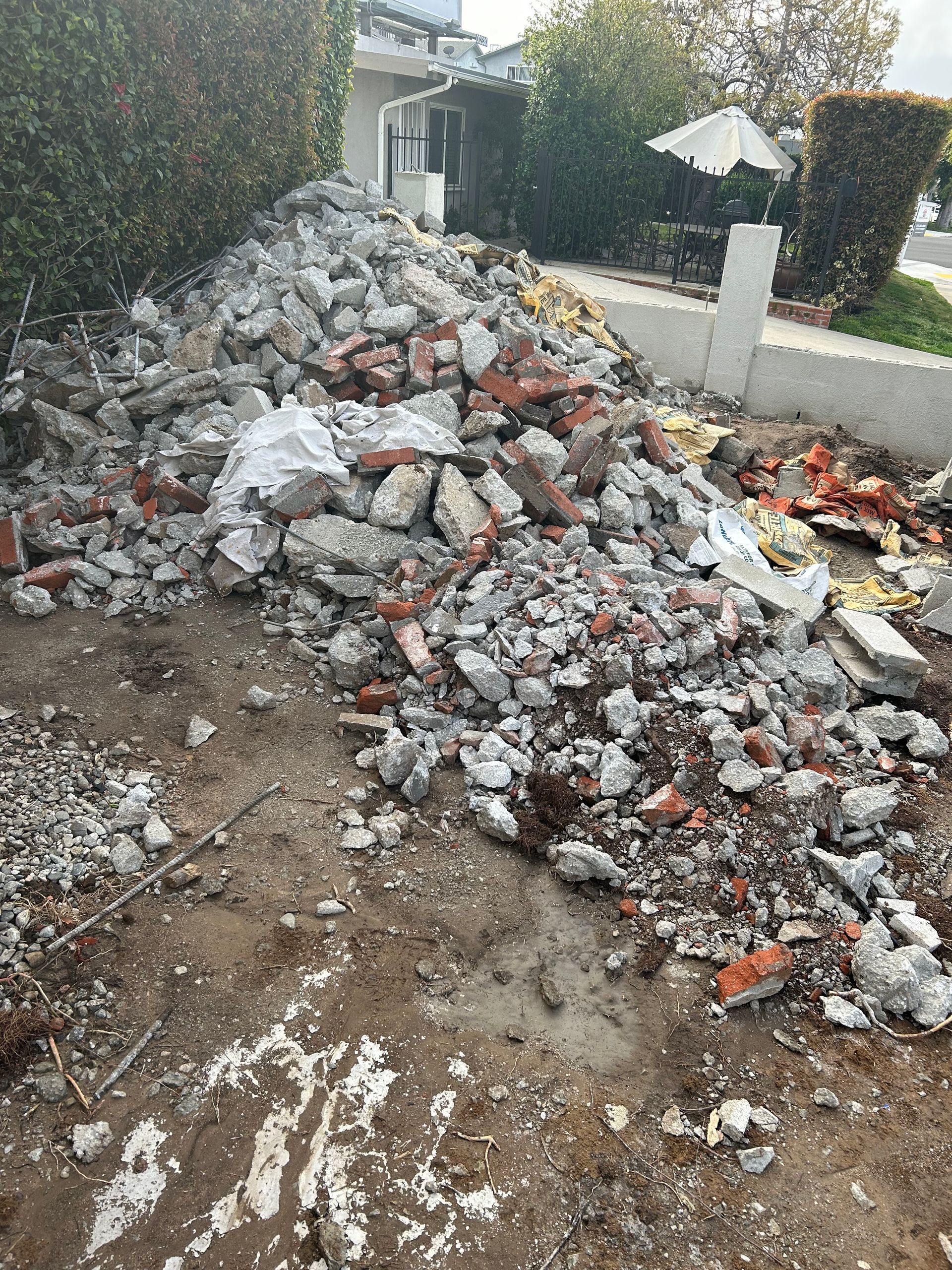 A large pile of broken concrete rubble and bricks sitting on dirt near a house and hedge.