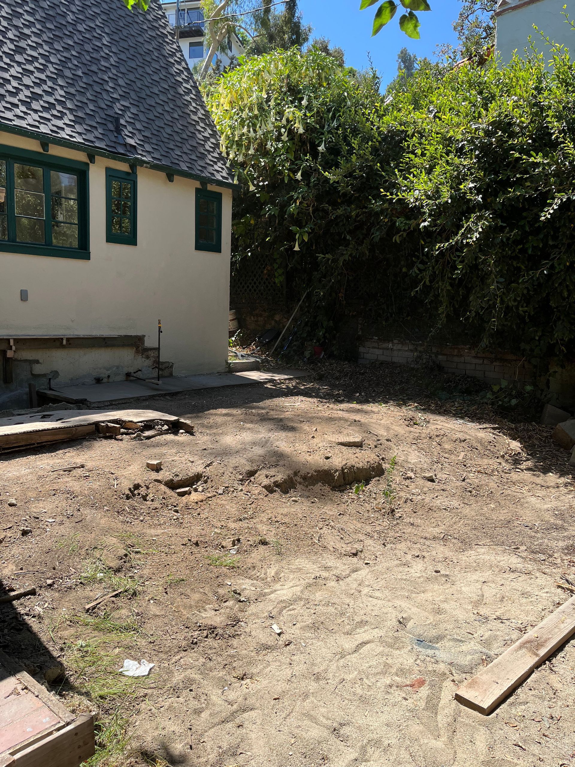 A cream-colored house with dark green trim overlooks a bare, sandy backyard bordered by dense green trees and shrubs.