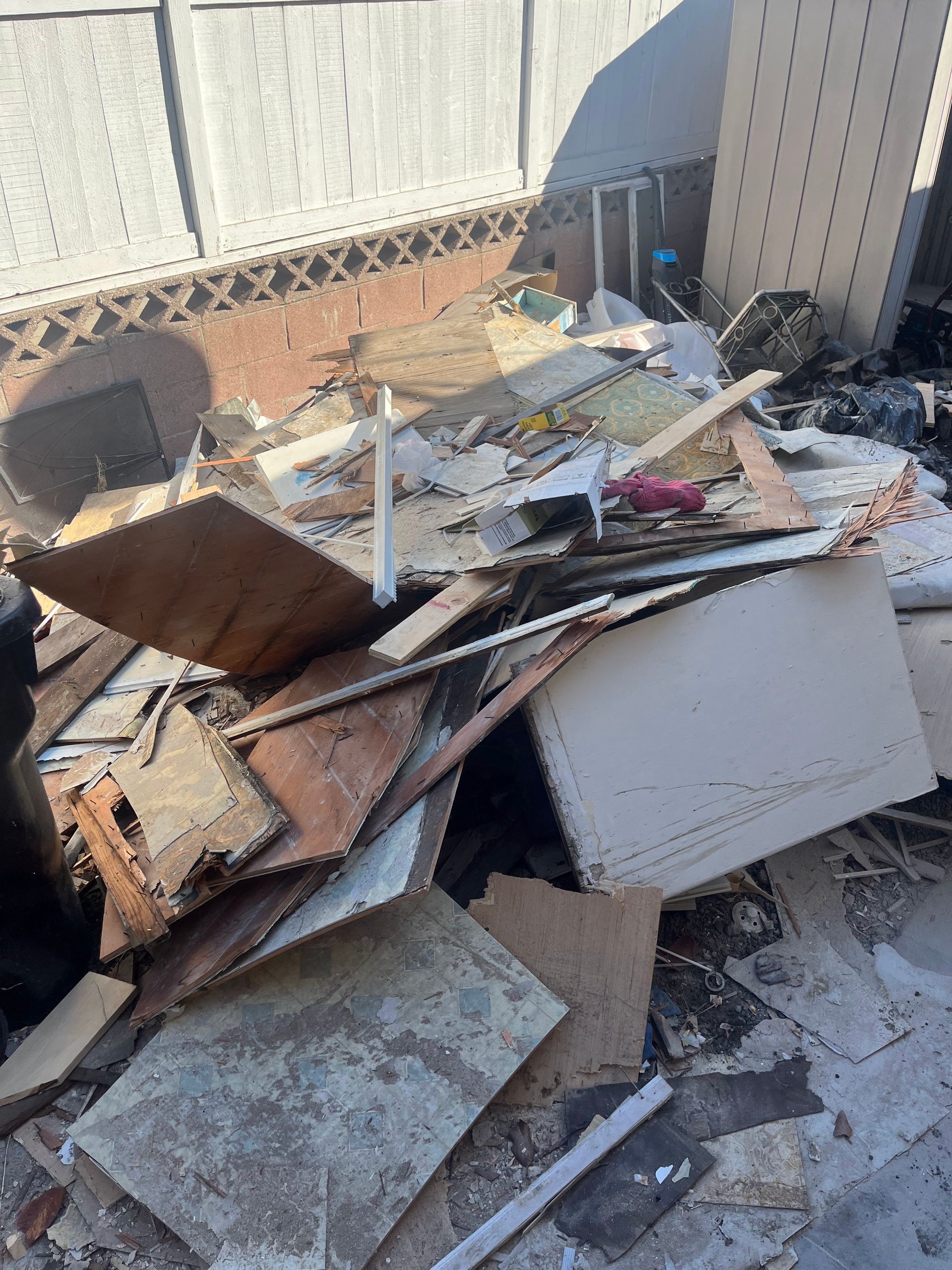 A pile of construction debris, including drywall, wood scraps, and rubble, sitting outside against a white fence.
