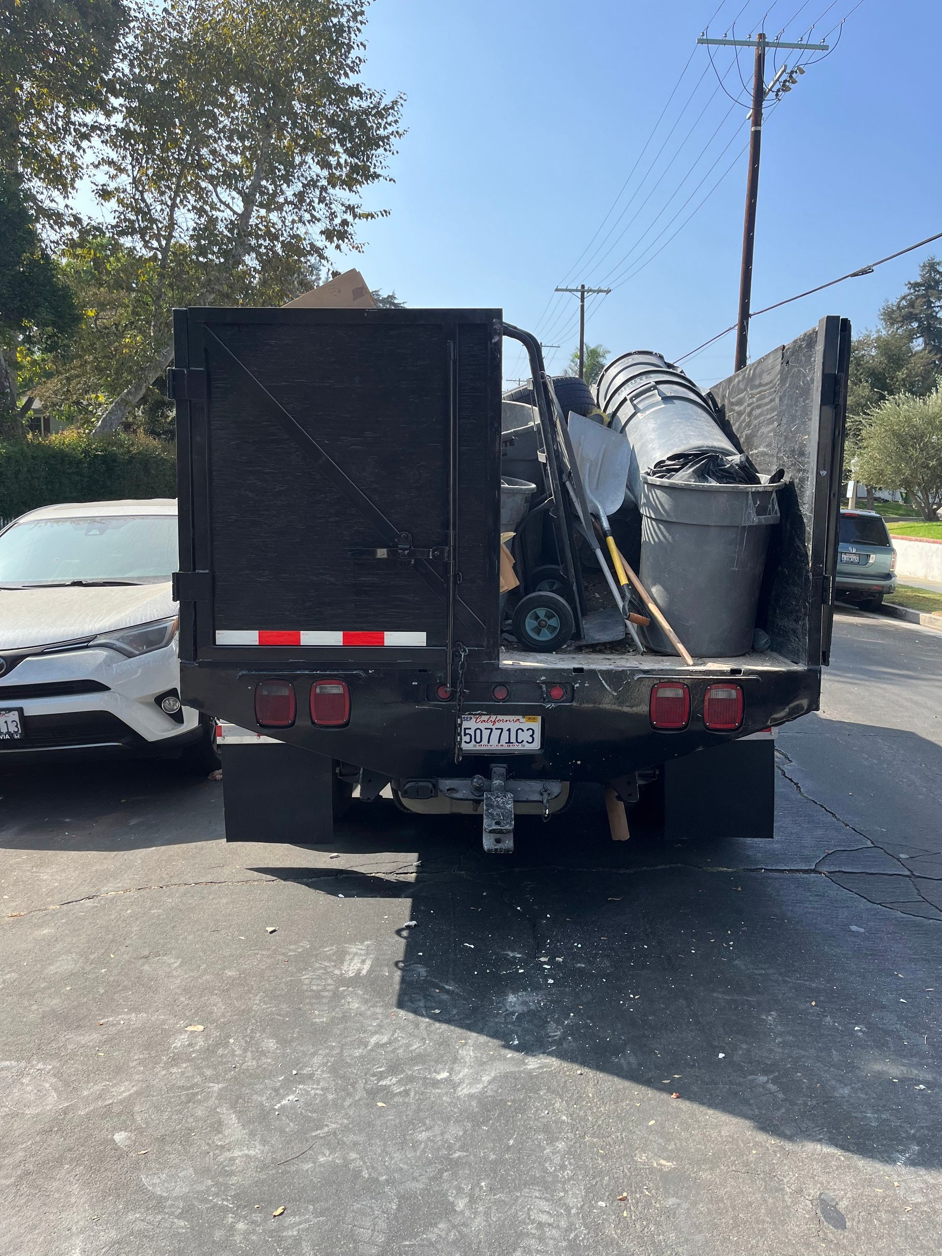 A black dump truck parked on a street, filled with gray trash cans and assorted debris, next to a white car.