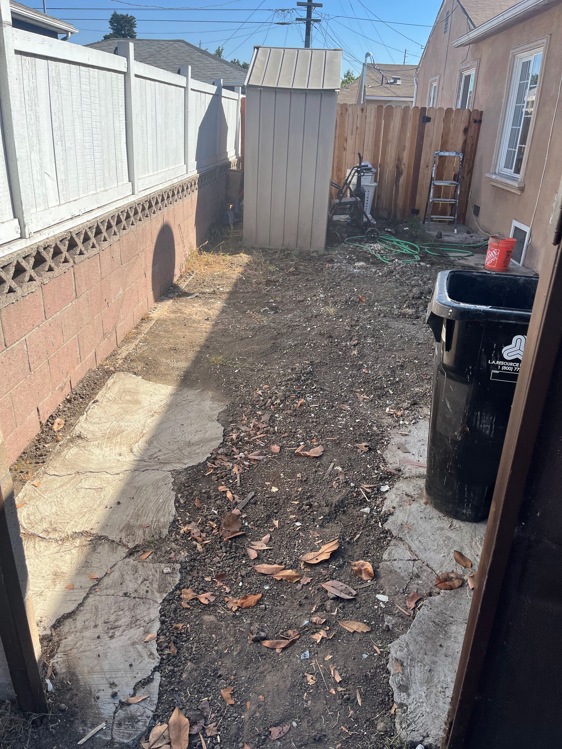 A narrow backyard pathway with cracked concrete, scattered leaves, a tall storage shed, and a black trash bin.