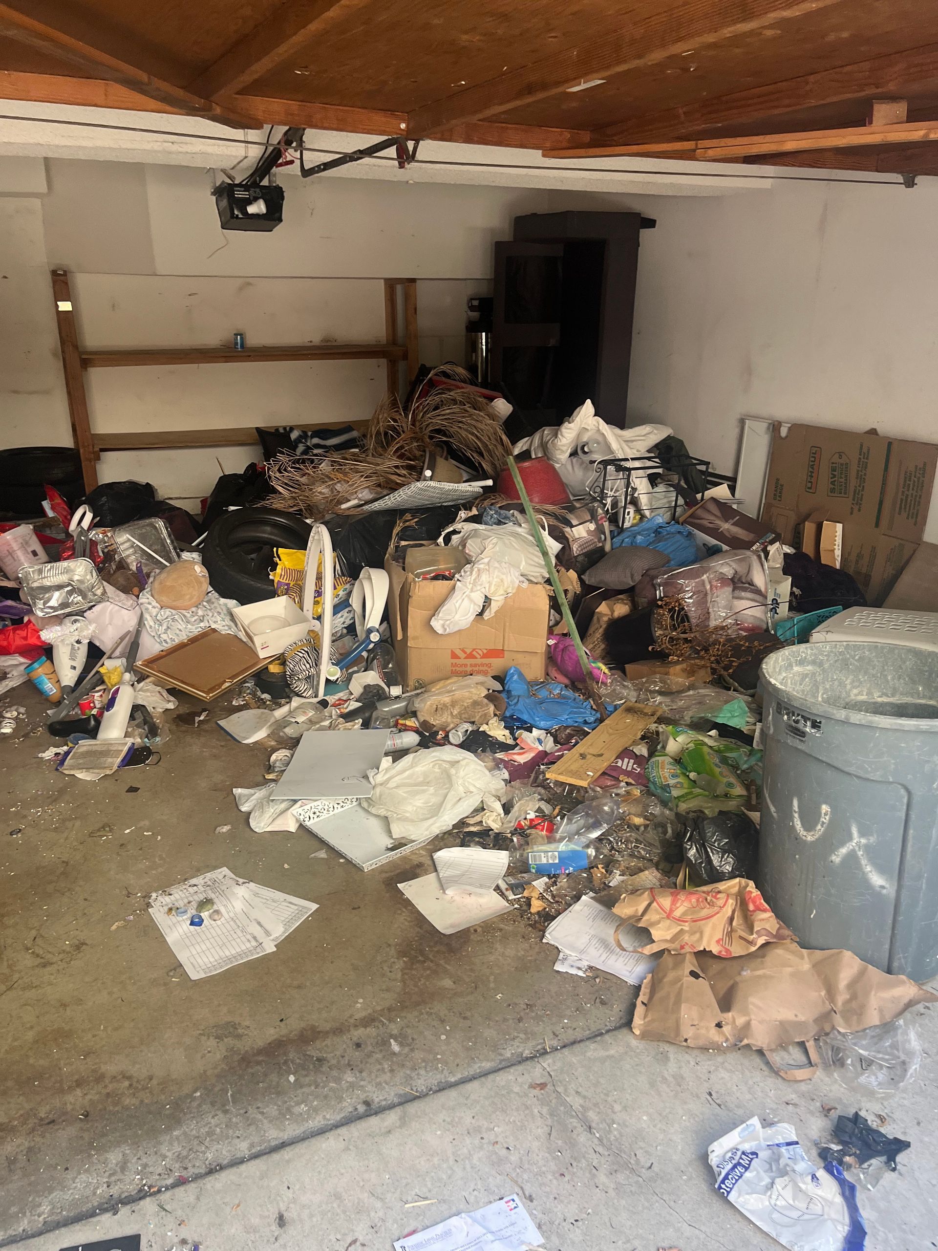 A cluttered garage space filled with scattered trash, cardboard boxes, and miscellaneous debris on a concrete floor.