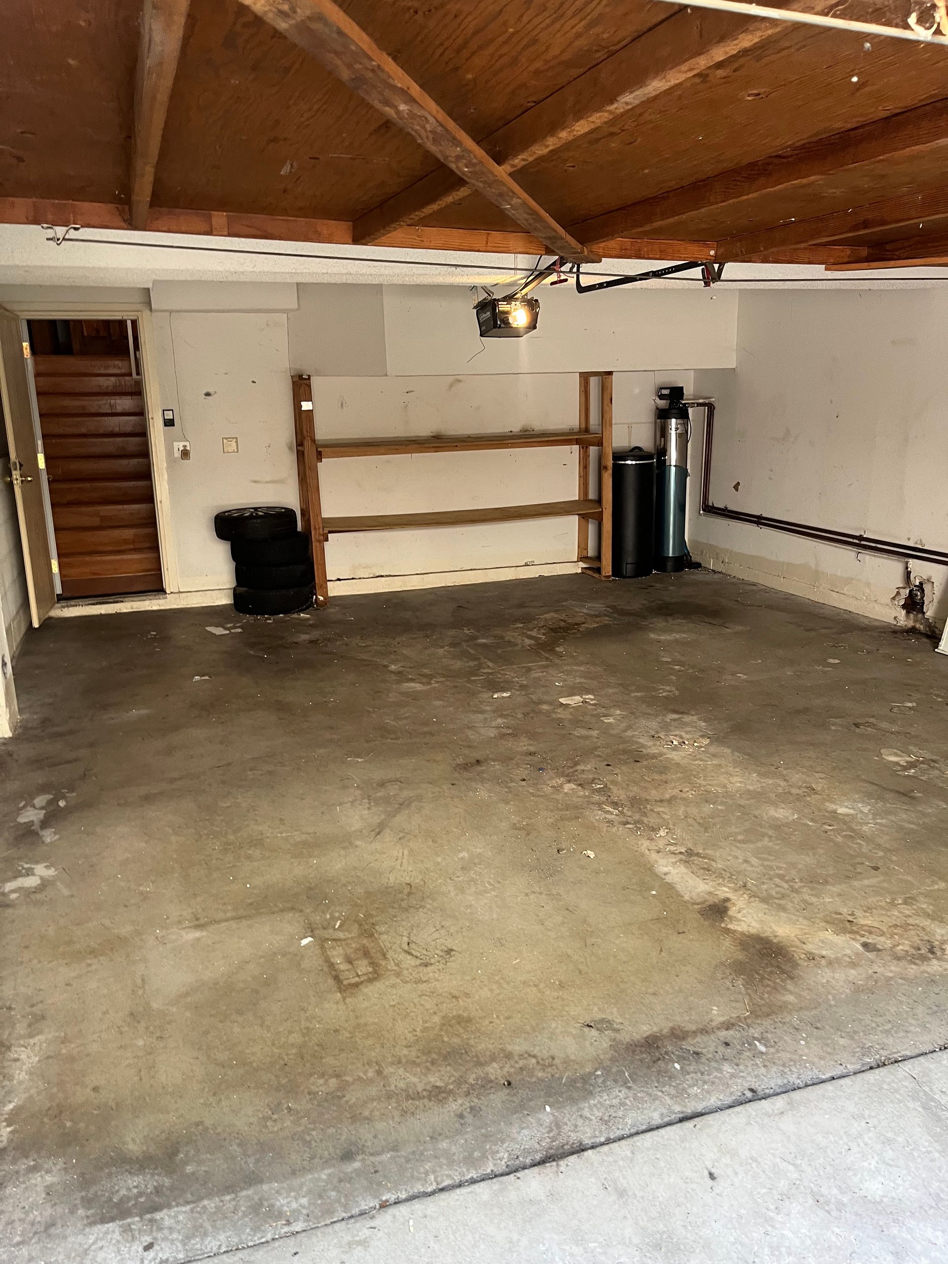 An empty garage with a concrete floor, wooden ceiling beams, wooden shelving, a set of stairs, and a water softener unit.