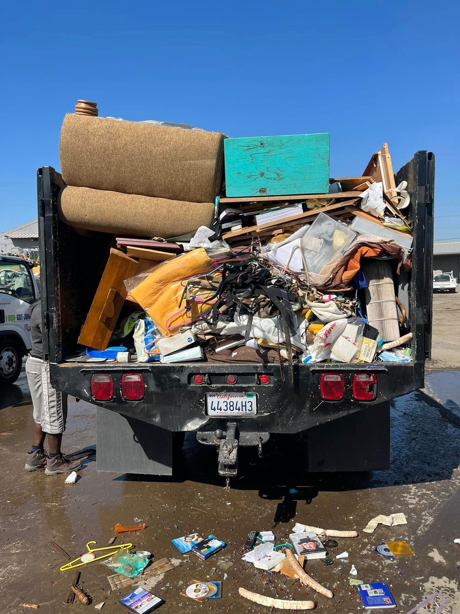 A truck bed piled high with assorted debris, including rolled carpet, a green container, and scattered litter on the ground.