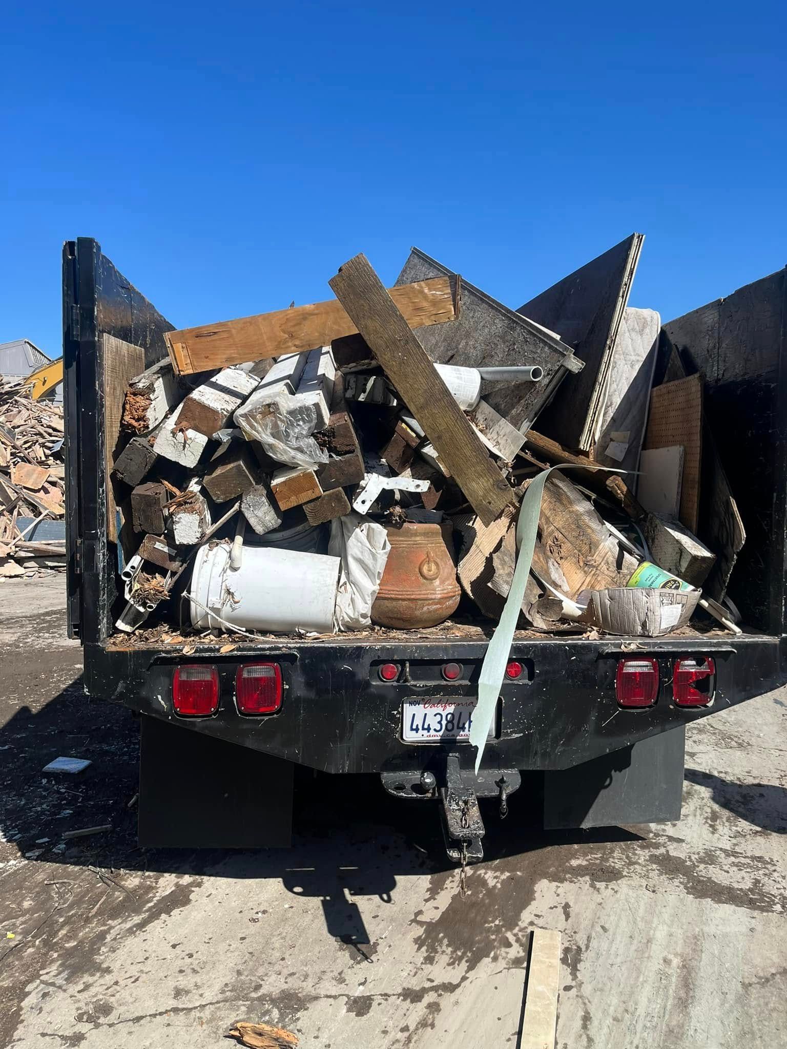 The back of a black truck filled with construction debris, lumber, and various household waste under a clear blue sky.