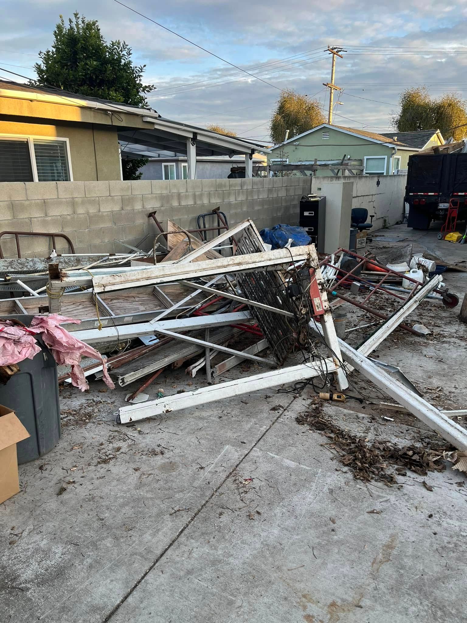 A collapsed white metal awning structure lies on a concrete driveway in front of a residential wall and houses.