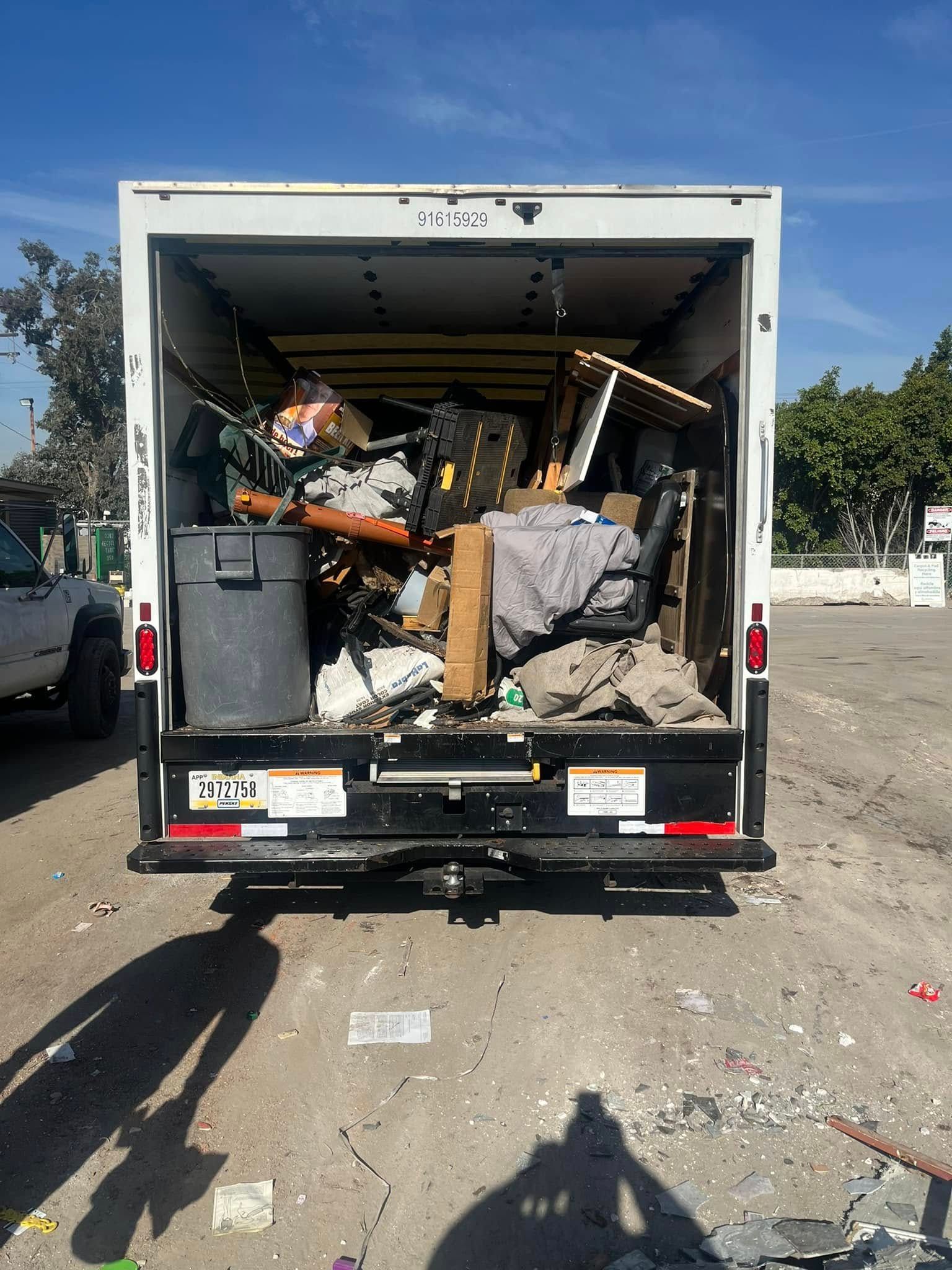 The open back of a white moving truck parked on a dirt lot, filled with various boxes, furniture, and a trash bin.