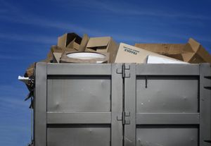 A gray industrial dumpster filled to the brim with discarded cardboard boxes against a bright blue sky.