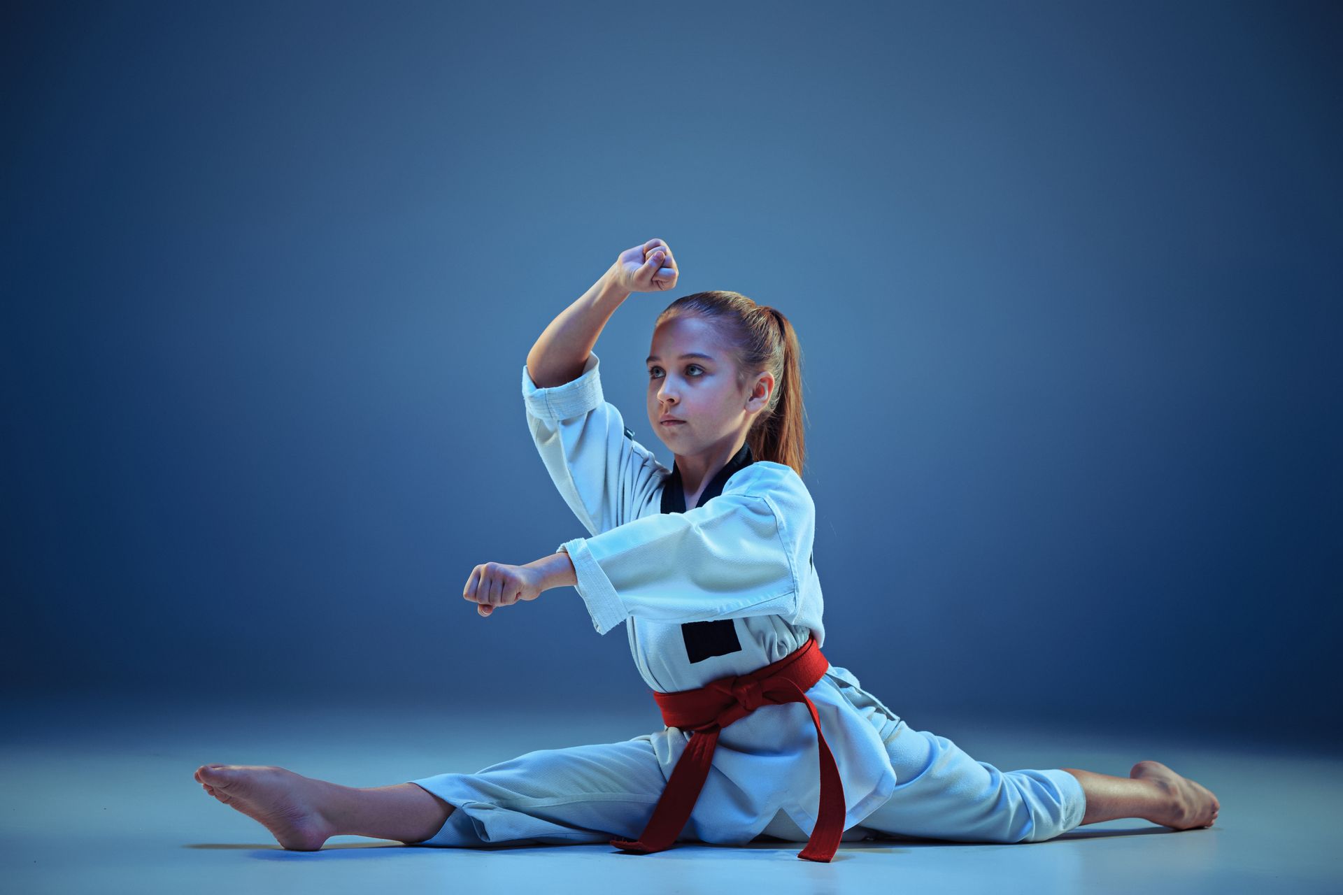 A young girl in a karate uniform is doing a split on the floor.