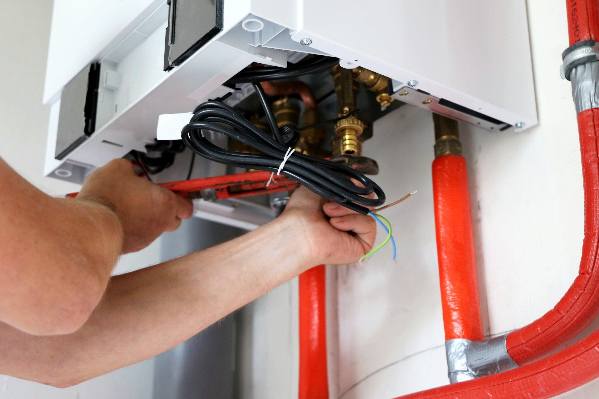 Hands working on wiring under a white appliance with red pipes and black cables in a utility space