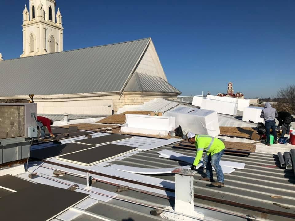Construction workers install roofing on a building with a white steeple under a clear blue sky.