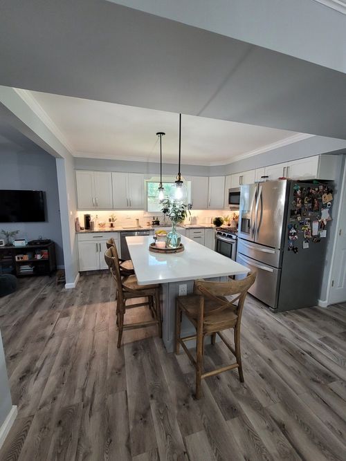 A modern kitchen featuring white cabinets, a large white island with two wooden stools, stainless appliances, and wood floors.