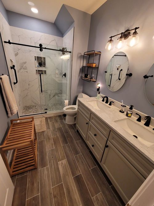 A bathroom featuring a gray vanity with double sinks, a glass-enclosed shower, a toilet, and dark wood-look tile flooring.