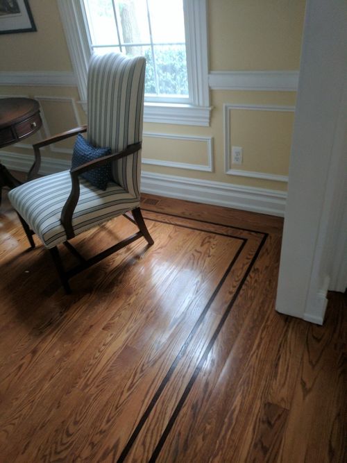 A striped armchair sits on hardwood flooring featuring a decorative dark border near a window with yellow wainscoted walls.