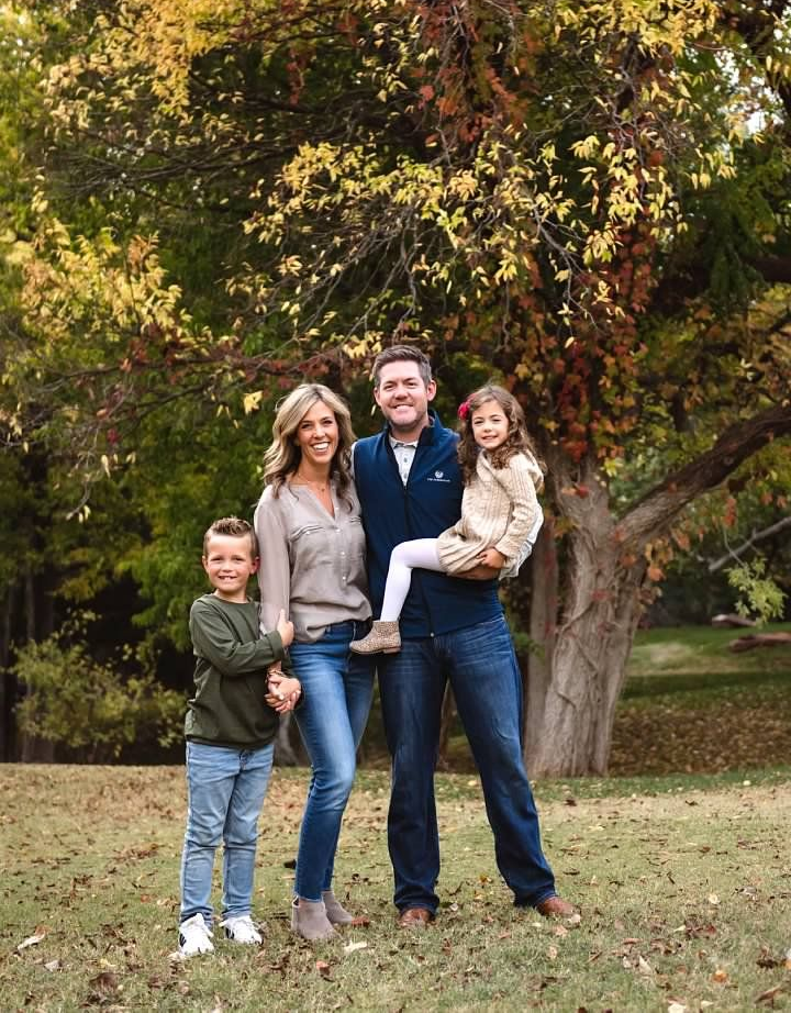 Family of four poses outdoors under a tree. Parents and two children smile, with autumn foliage in the background.