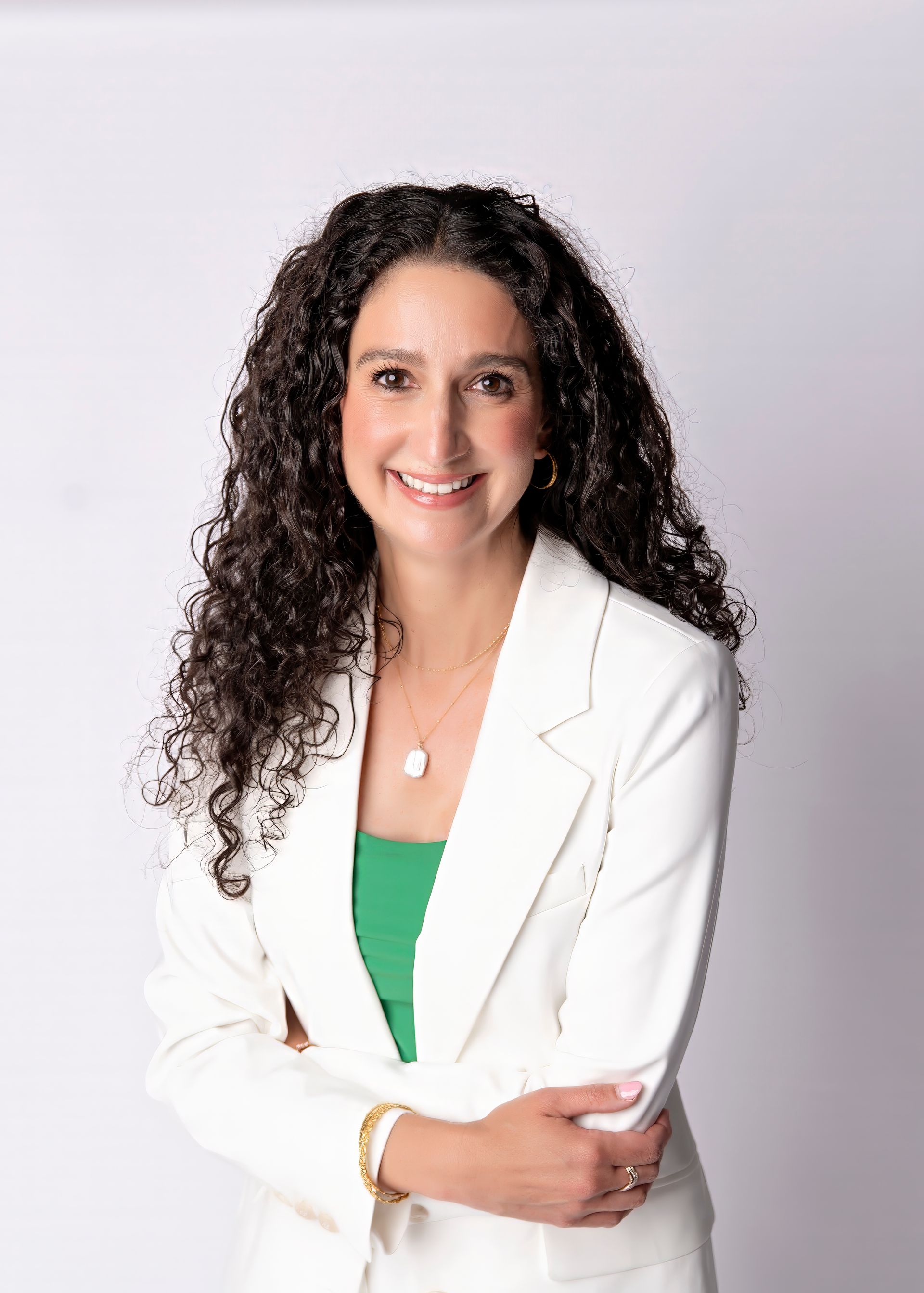 Woman with dark curly hair smiles, wearing a white blazer over a green top. She has a gold bracelet and a necklace.
